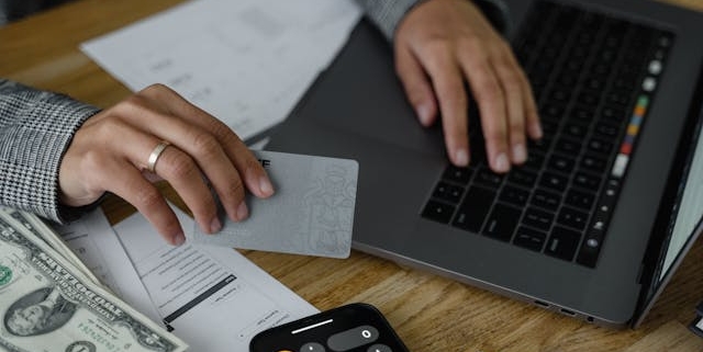 person-using-a-laptop-while-holding-a-bank-card-with-cash-documents-and-a-calculator-on-the-table person-using-a-laptop-while-holding-a-bank-card-with-cash-documents-and-a-calculator-on-the-table