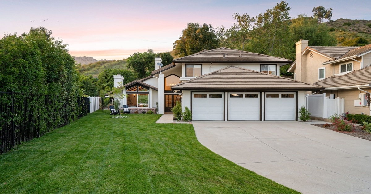 Two-story suburban house with three-car garage, manicured lawn, patio seating, and surrounding trees under a pink and blue evening sky—ideal for luxury property management in Salt Lake City. Residential and luxury property management home in Salt Lake City Utah
